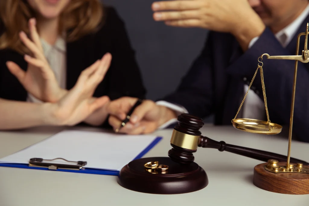 Two people sit at a table with a document. A gavel, balance scales, and two rings are on the table, suggesting a legal or divorce discussion.