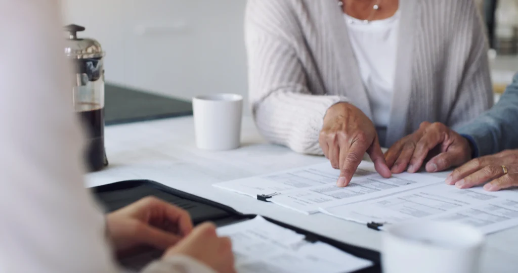 Two people sitting at a table, one pointing at documents. A coffee cup and a clipboard are visible on the table.