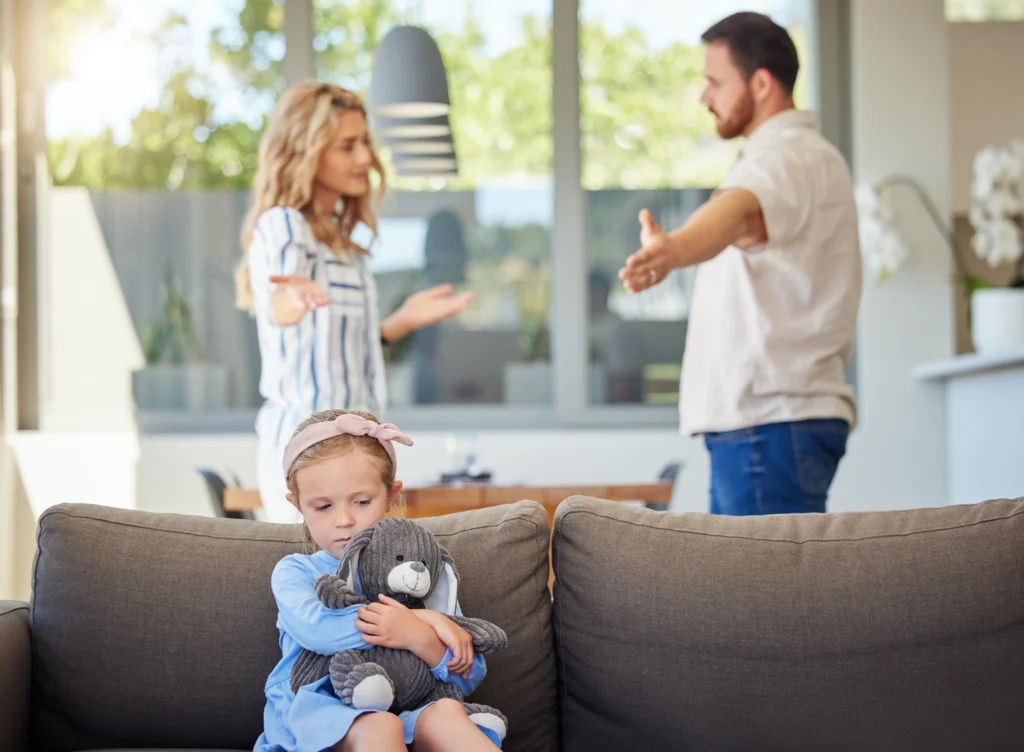 A young girl sits on a couch hugging a stuffed bear, looking concerned, while a man and woman in the background appear to be arguing.
