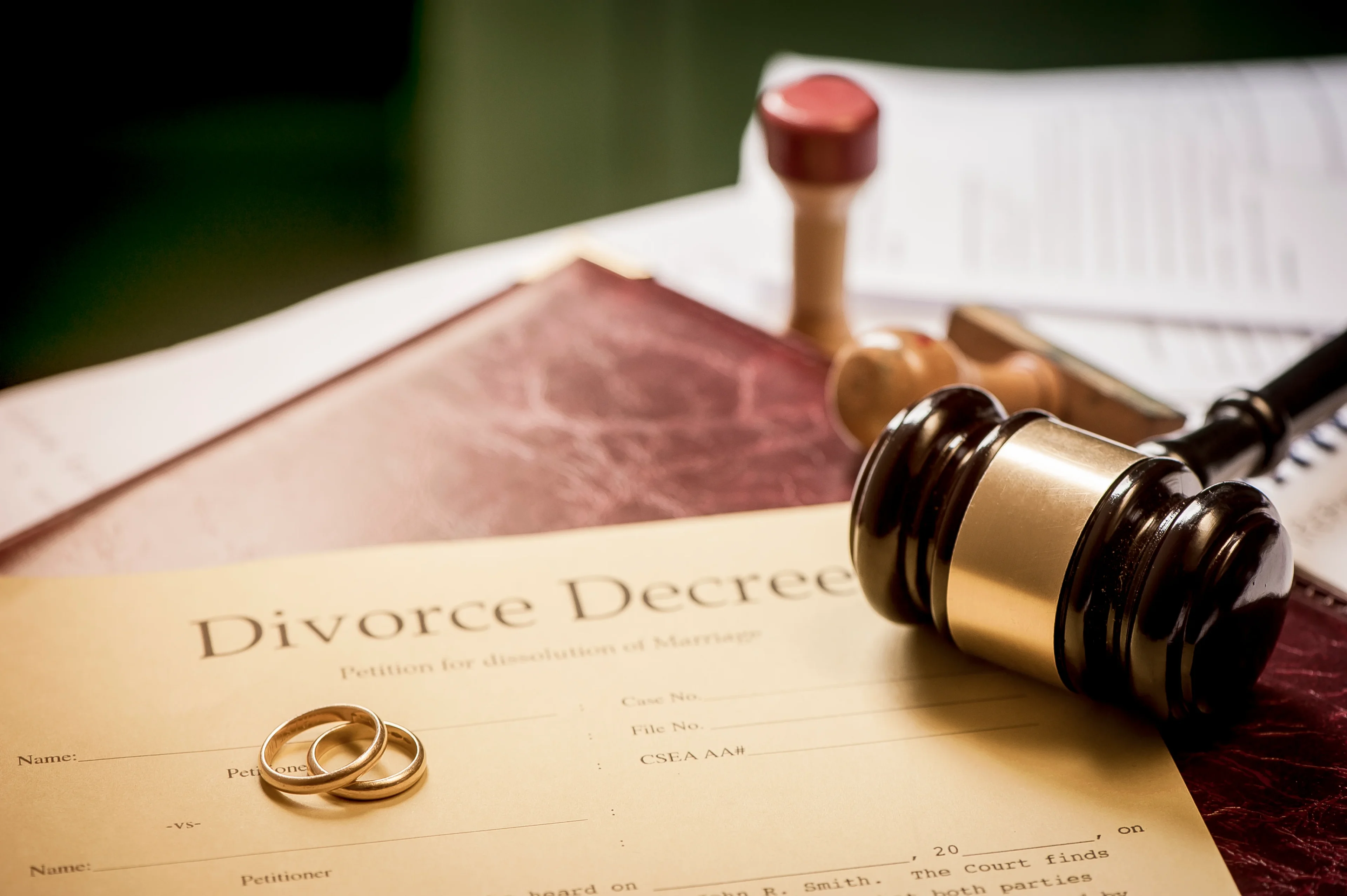 Close-up of a divorce decree with two wedding rings on top. A judge's gavel, stamps, and some paperwork are visible in the background on a desk.