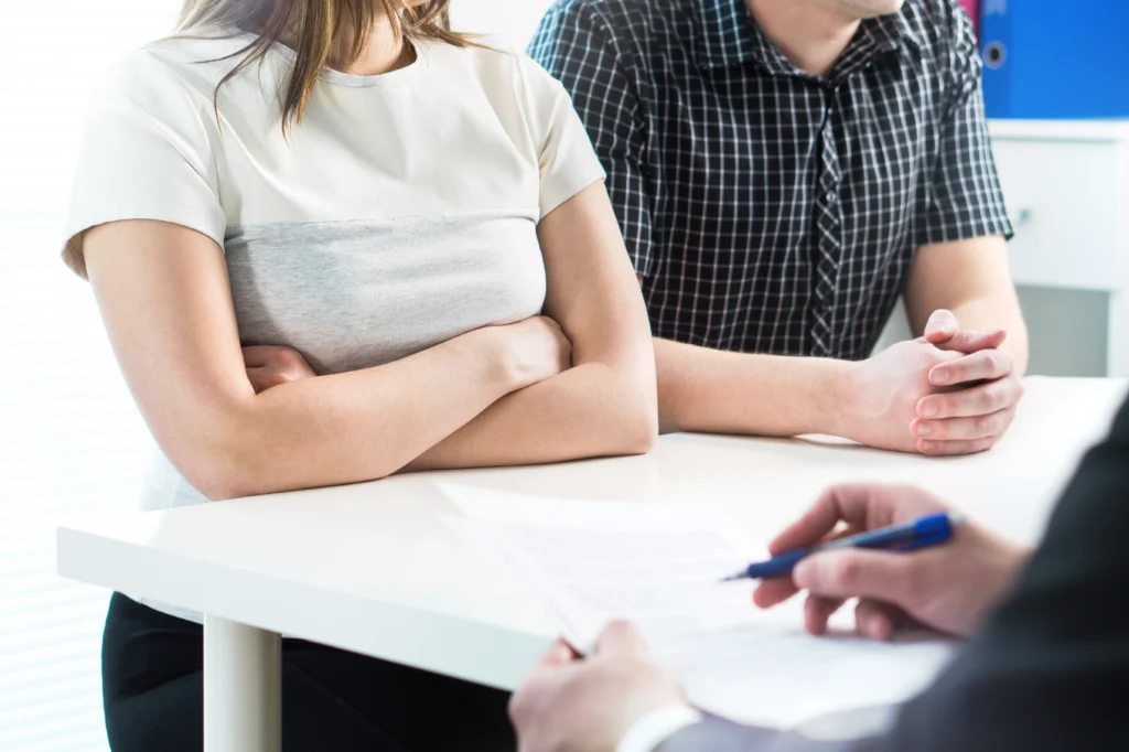 Two people sit across from a person holding a pen and paper, indicating a discussion or meeting. The woman has her arms crossed on the table.